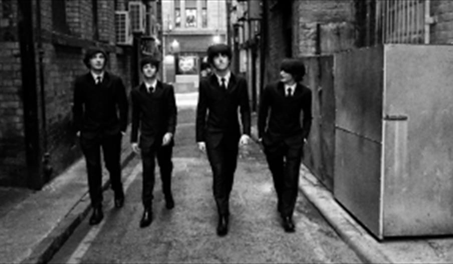A black and white photo of 4 men dressed as the Beatles walking down an alleyway