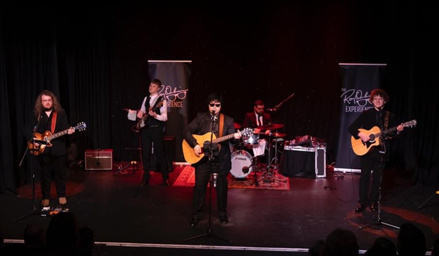 A band performs energetically on stage with guitars, bass, and drums. Members wear black suits, creating a classic rock ambiance. Two banners flank th