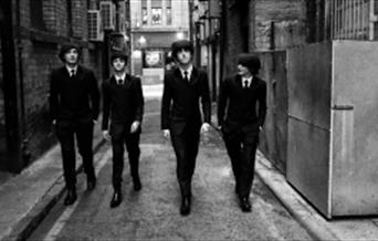 A black and white photo of 4 men dressed as the Beatles walking down an alleyway