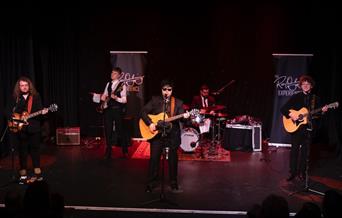 A band performs energetically on stage with guitars, bass, and drums. Members wear black suits, creating a classic rock ambiance. Two banners flank th