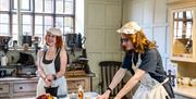 Two people dressed in aprons and bonnets, pose in a Victorian styled kitchen