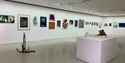 white gallery space with artworks in a line on the wall, on the floor is a a sculpture shaped like an anchor and a large white square plinth with smal
