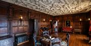 A large wood panel walled room with period features. The ceiling has an ornate relief design. The room is decorated with period furnishings including