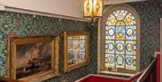The stairwell at Elizabethan House, showing a large stained glass window and walls decorated in a floral themed wallpaper. On the wall is a large pain