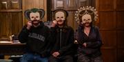 Three people sat in a room with wood panelled walls. Each visitor is holding an Elizabethan styled mask in front of their faces.
