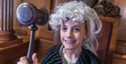 A young boy sits in a judges seat. He is wearing a judges wig, cape and is holding a gavel